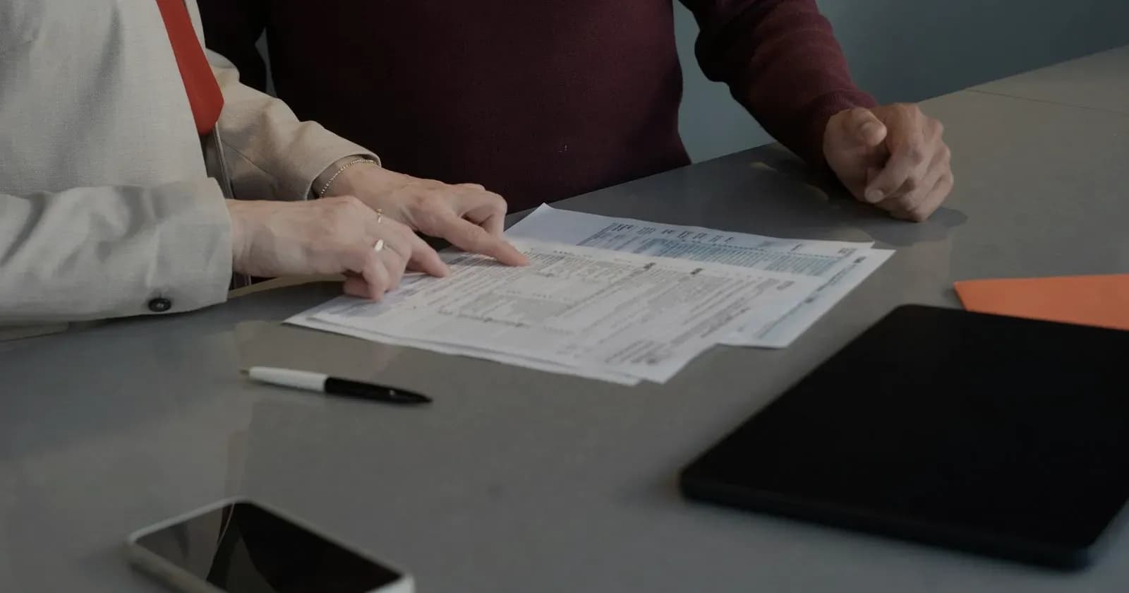 Two people seated across an office desk, one pointing out a line on a printed statement between them.