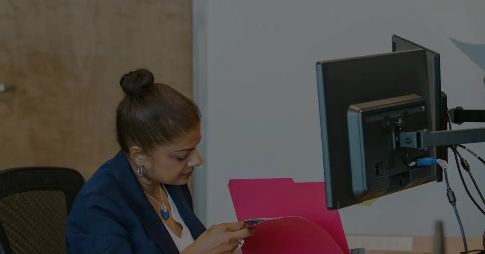 A professional in profile at a desk, opening a client file folder beside a monitor.
